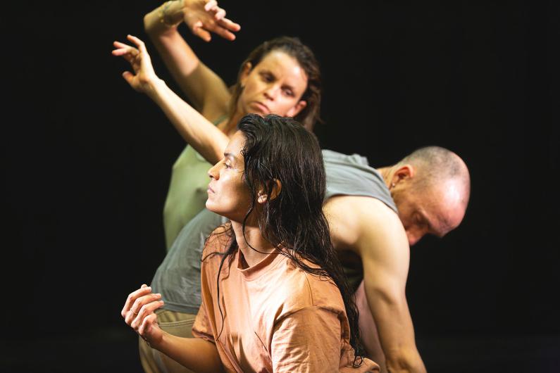 Drei Personen bewegen sich in einer Tanzperformance auf einer dunklen Bühne. Eine Person im Vordergrund trägt ein lockeres T-Shirt und blickt konzentriert zur Seite. Zwei weitere Personen im Hintergrund bewegen synchron ihre Arme.
