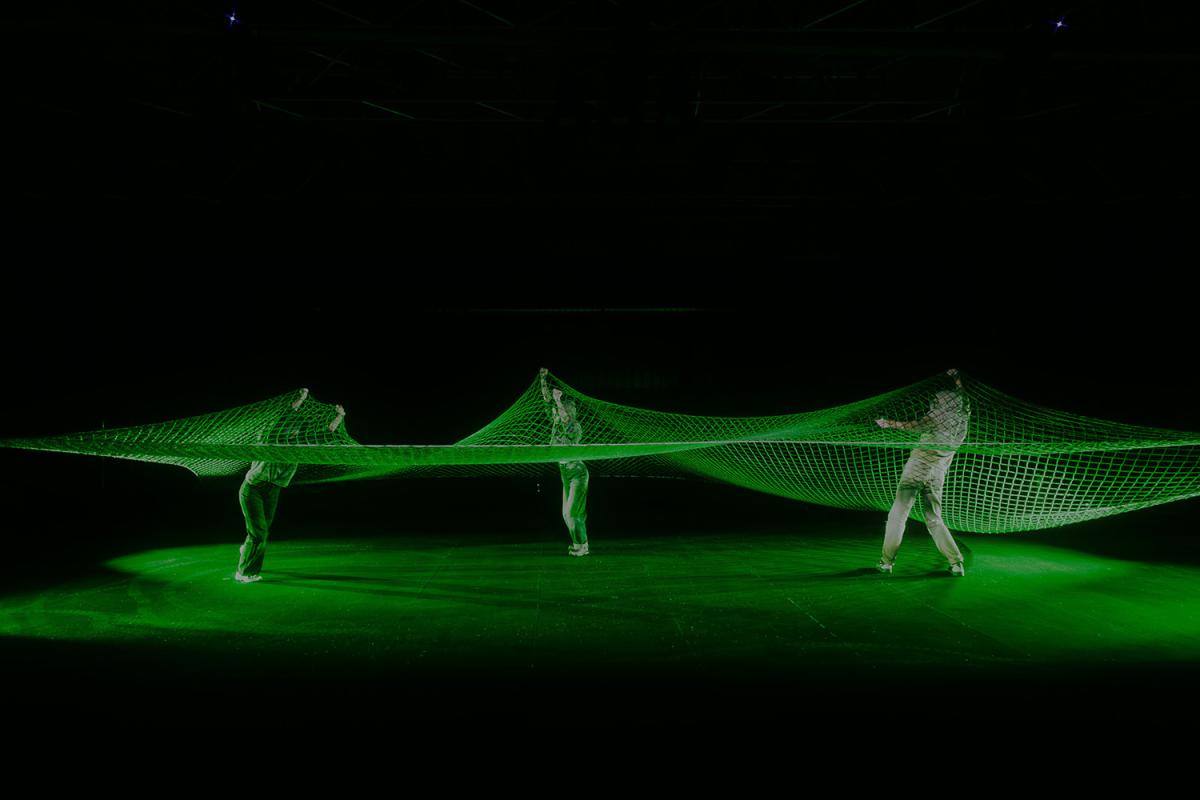 Three people stretch a large green net across a stage. The net forms a curved line, and the scene is bathed in green light.