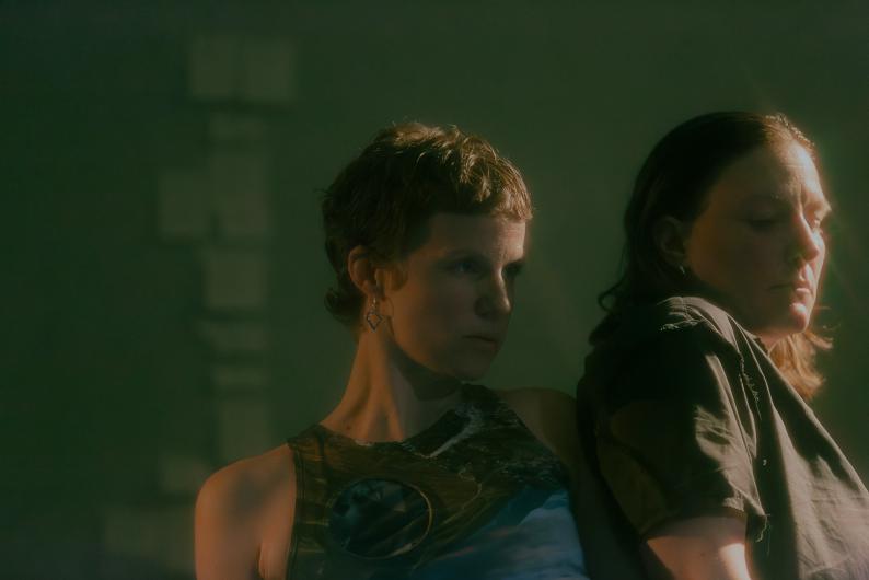 Two people rest their shoulders against each other, calm and focused. Soft light falls on their faces, while the background remains dark green.