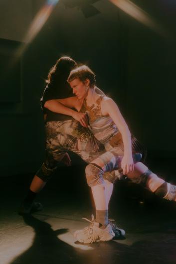 Two people stand close together in a dance pose, one leaning over the other. Warm stage light highlights their movement