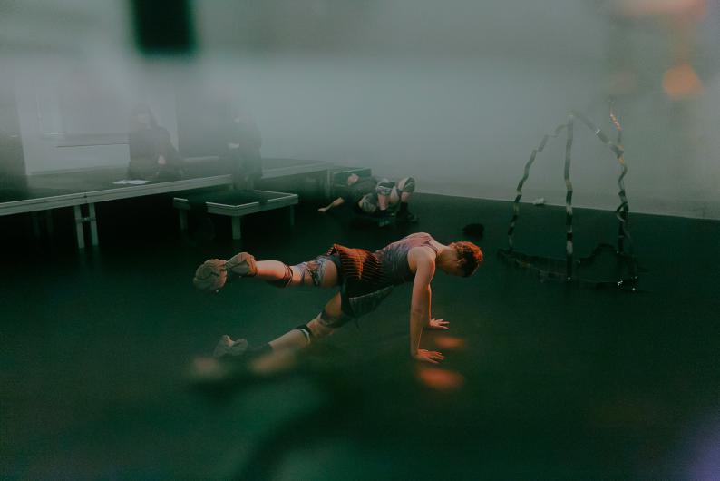 A person supports themselves on hands and feet on the floor, while another lies in the background. Mist and soft light create a diffuse atmosphere.