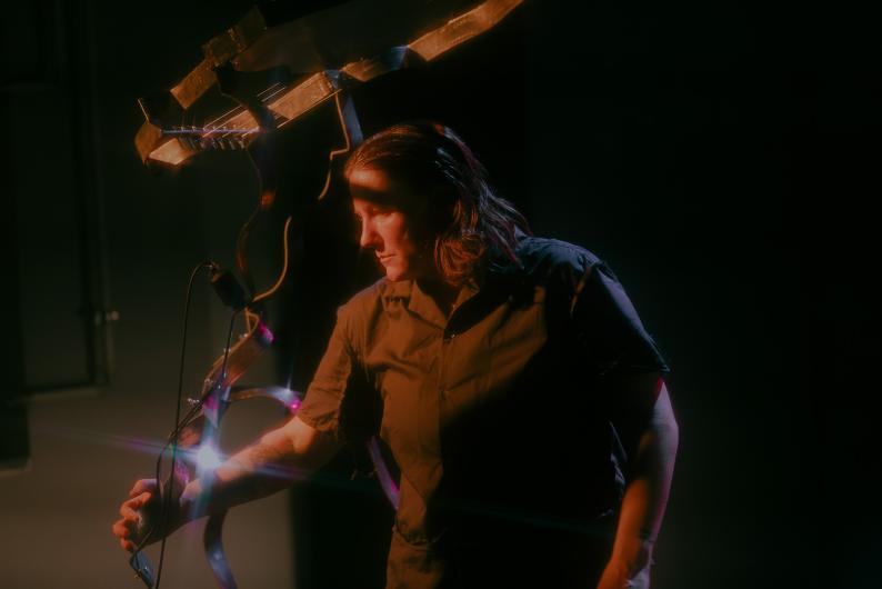 A person stands in semi-darkness next to a metal sculpture made of curved strips. The light highlights their face and the structure.