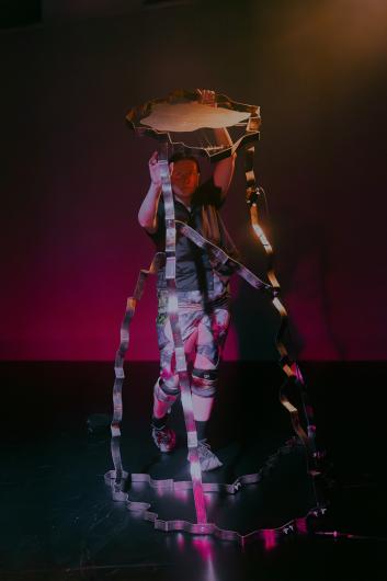 A person holds a large, wavy metal sculpture above their head, illuminated by stage light.