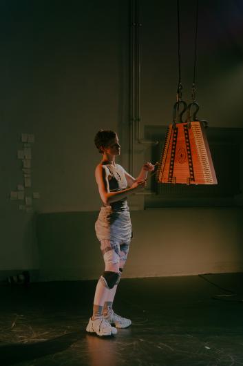 Two people face each other on a dimly lit stage, each illuminated with their own object – a metal structure on the left, a wooden object on the right.