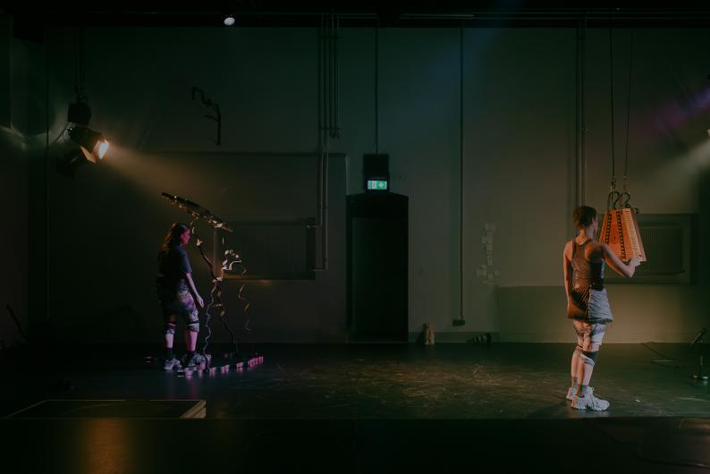 A person kneels on the floor beneath a large hanging wooden instrument, moving in a dance-like pose.
