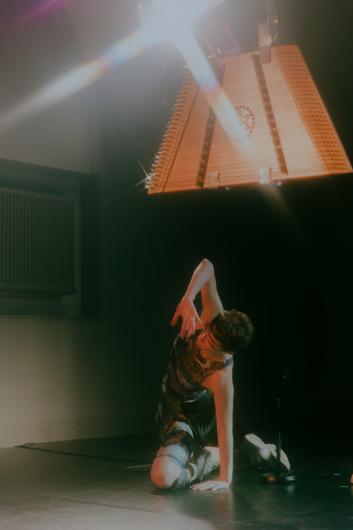 Eine Person steht auf der Bühne, umgeben von sanftem Licht, und hält eine geschwungene Metallstruktur. Die Atmosphäre ist ruhig und konzentriert.