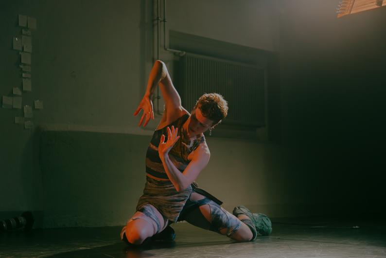 A person kneels on stage, moving their arms and hands in an expressive dance pose. Warm light shines from above, with a dark background.