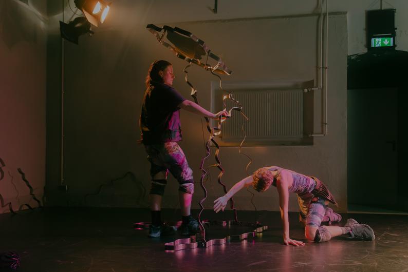 One person stands next to a wavy metal sculpture, holding it, while another kneels in front, reaching out with an extended hand. The scene is bathed in soft reddish stage light.