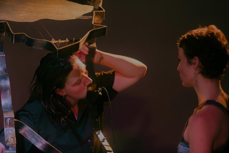 Two people face each other on stage. One holds a metal sculpture above their head, looking at it intently, while the other watches attentively. Warm light highlights their faces and the structure.