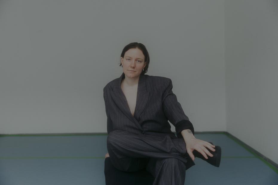 A person sits on the floor in an empty room. They are wearing a dark pinstripe suit, with one leg crossed, looking calmly at the camera.
