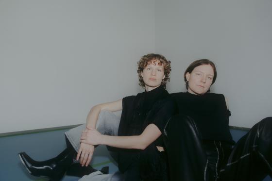 Two people are sitting relaxed next to each other in a simple, bright room. Both are wearing dark tops and are looking calmly and directly at the camera.