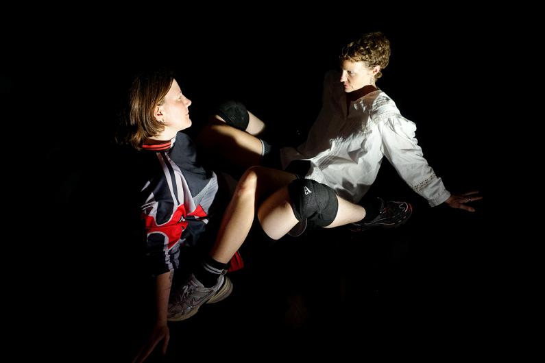Two people sit facing each other on the floor, lit by a sharp beam of light, wearing knee pads and looking at each other intensely.