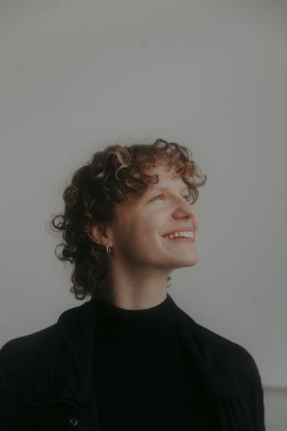 A person with curly hair smiles and looks upward. They are wearing a dark top, with a plain light background.