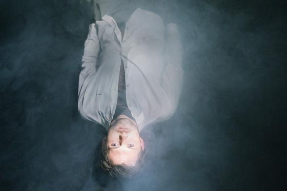 A person lies in dense fog or smoke. They wear a light shirt and look upward, illuminated from above.