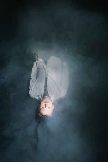 Eine Person liegt in dichter Nebel- oder Rauchkulisse. Sie trägt ein helles Hemd und blickt nach oben, während sie von oben beleuchtet wird.
