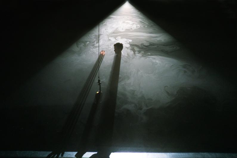 Eine Person steht in einem Lichtstrahl, umgeben von Nebel. Ein Arm ist erhoben und berührt den Lichtkegel, während lange Schatten entstehen. Ein Mikrofon hängt von der Decke herab.