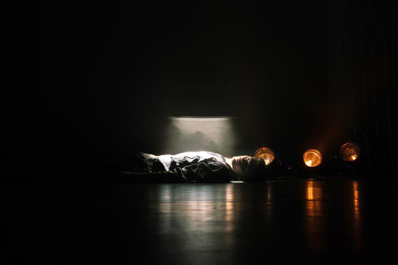 Eine Person liegt auf dem Boden in einem dunklen Raum. Ihr Gesicht und Oberkörper werden von einem schmalen Lichtstrahl und Bühnenlichtern im Hintergrund beleuchtet.