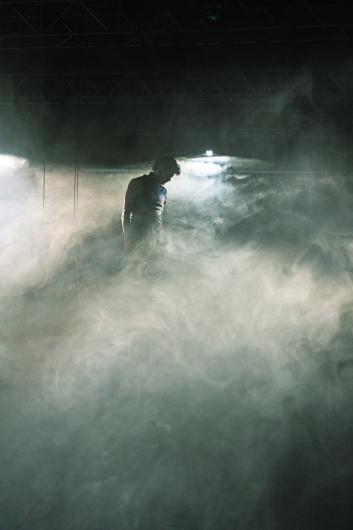 Eine Person sitzt auf einem Stuhl auf einer leeren Bühne und wird von Nebel umgeben. Über ihr sind Scheinwerfer und Bühnenstrukturen sichtbar, während eine Geige an einem Seil hängt.