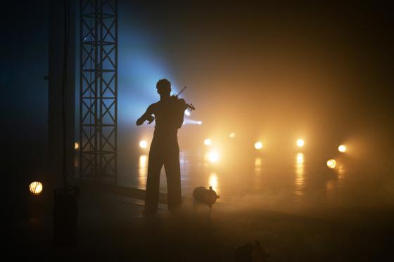 A person sits sideways in stage fog, holding a violin. Warm lights in the background create a dense, golden atmosphere.