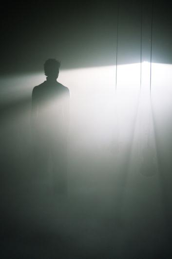 Eine Geige hängt im Vordergrund an einem Seil, während eine Person im Hintergrund im Lichtnebel sitzt. Die Szene ist in weiches, diffuses Licht getaucht.