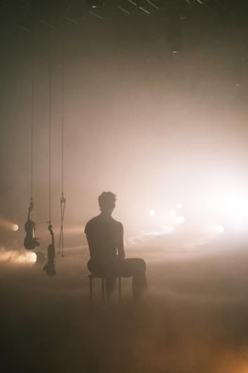 Eine Person steht im Lichtkegel und spricht oder singt in ein Mikrofon. Das Gesicht ist von sanftem Bühnenlicht erhellt, der Hintergrund bleibt im Nebel verborgen.