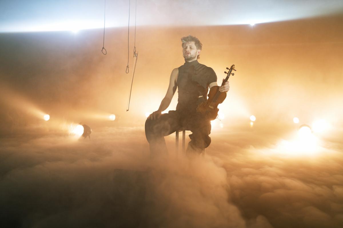 A person sits on a stool surrounded by dense fog, playing the violin. The stage is bathed in warm golden light, with numerous spotlights arranged around them.