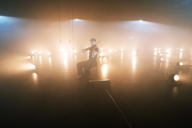 Eine Person sitzt auf einem Hocker in einem Raum voller Nebel und hält eine Geige in der Hand. Der Hintergrund ist in goldenes Licht getaucht, über der Szene hängen Seile und Lichtquellen.
