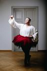 Eine Künstlerperson mit ausdrucksstarkem Make-up, gekleidet in eine weiße fließende Bluse und einen roten Tüll-Tutu, posiert in einer dramatischen Ballett-Haltung vor einer grauen Holztür. Die Hand ist anmutig ausgestreckt, der Blick intensiv.