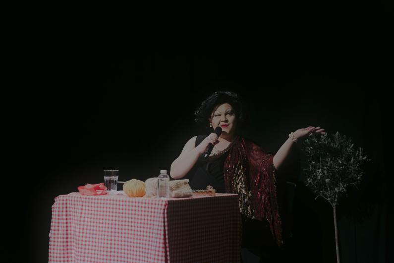 Person sitting at a table with a red-and-white checkered tablecloth, speaking into a microphone, with water and yarn beside them.