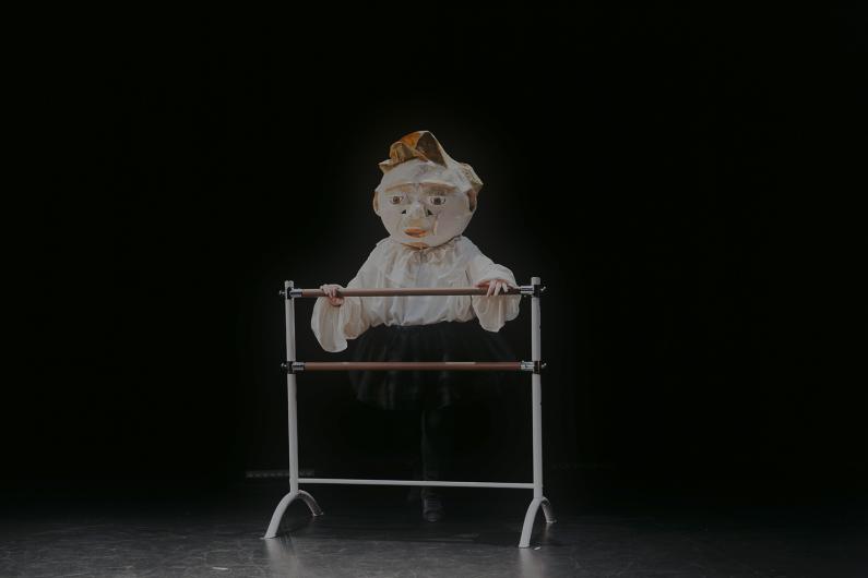 Person mit einer großen Papiermaske steht an einer Ballettstange, in einem dunklen Raum beleuchtet.