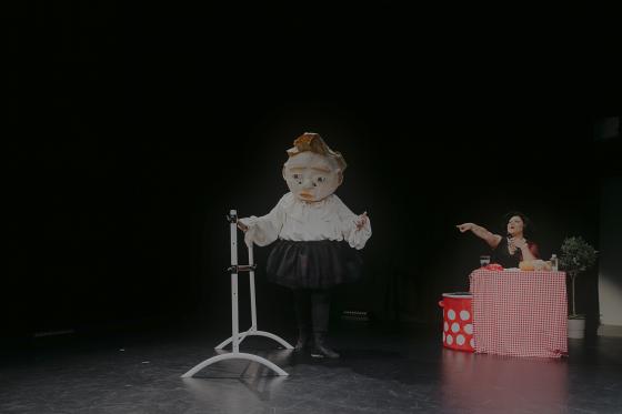 Maskierte Figur steht neben Tisch mit rot-weißer Tischdecke, während eine Person auf sie zeigt.