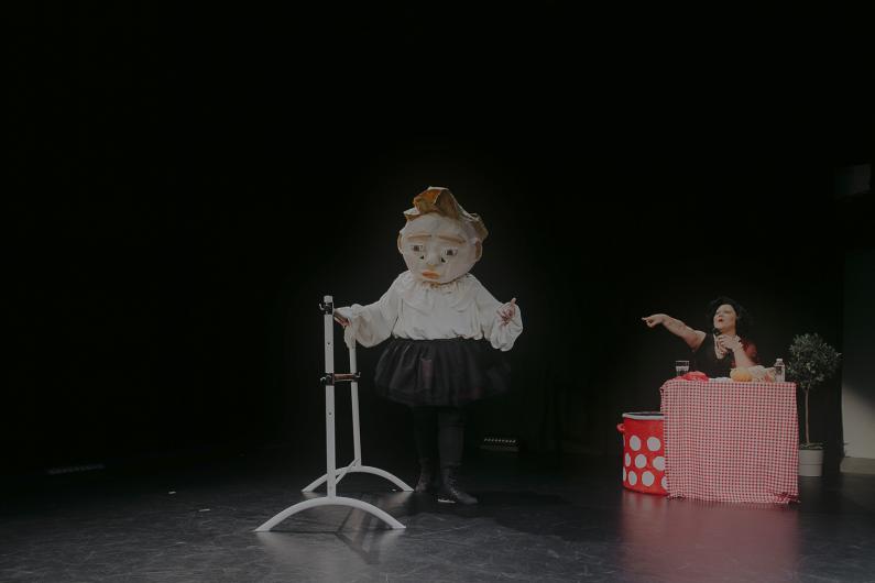 Masked figure stands next to a red-and-white checkered table, while a person points at them.