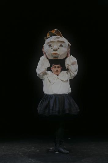 Person hebt eine große Papiermaske über ihren Kopf, trägt ein weißes Hemd und einen schwarzen Rock.
