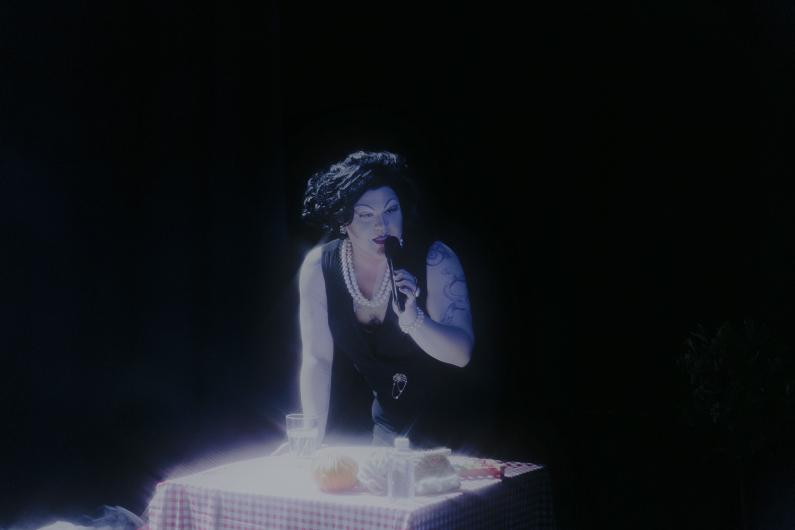 Person leans over a red-and-white tablecloth-covered table with a microphone, illuminated by dramatic lighting.