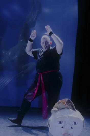 Person with a red sash strikes a theatrical pose on stage next to a large papier-mâché mask.