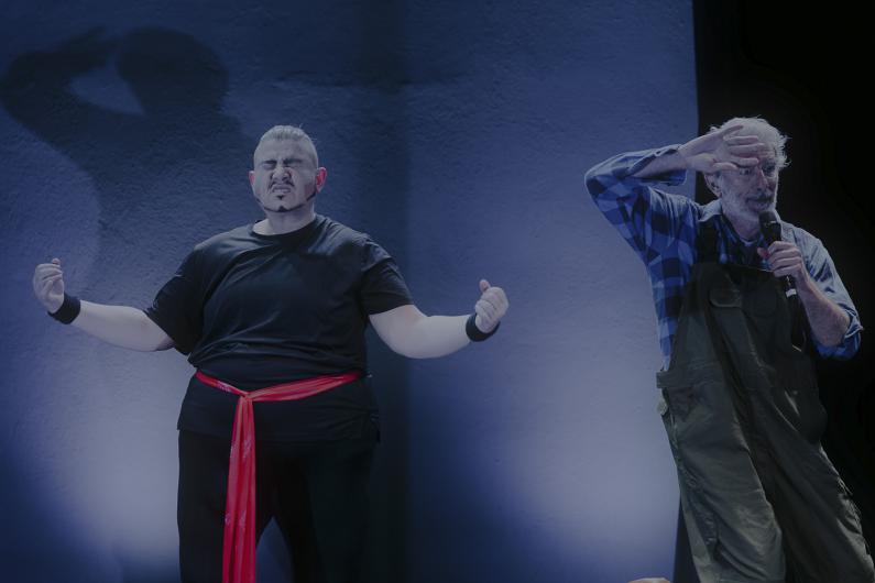 Zwei Performer auf der Bühne: eine mit zusammengekniffenen Augen und rotem Gürtel, der andere spricht ins Mikrofon.