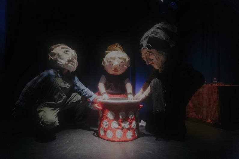 Drei Figuren mit großen Papiermasken knien um einen rot-weiß gepunkteten Behälter.