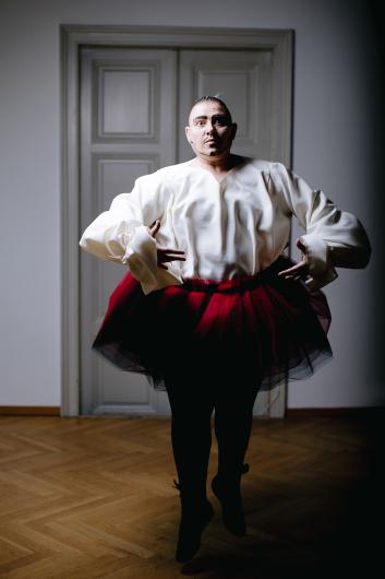 Eine Künstlerperson in einer weißen, fließenden Bluse und einem roten Tüll-Tutu nimmt eine elegante Ballett-Pose ein. Ihr intensiver Blick und das theatralische Make-up verstärken die dramatische Atmosphäre, während der Holzboden und die graue Tür eine klassische Kulisse schaffen.