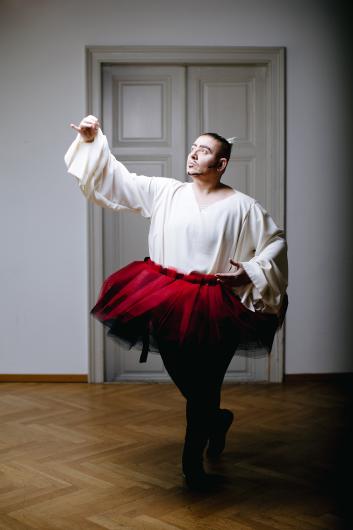Eine Künstlerperson mit ausdrucksstarkem Make-up, gekleidet in eine weiße fließende Bluse und einen roten Tüll-Tutu, posiert in einer dramatischen Ballett-Haltung vor einer grauen Holztür. Die Hand ist anmutig ausgestreckt, der Blick intensiv.