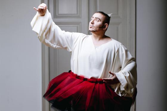 A performer with expressive makeup, wearing a white flowing blouse and a red tulle tutu, poses in a dramatic ballet stance in front of a gray wooden door. Their hand is extended gracefully, and their gaze is intense.