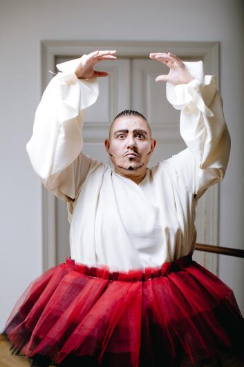 Eine Künstlerperson in einer weißen Bluse mit weiten Ärmeln und einem roten Tutu steht vor einer grauen Tür und hebt beide Hände in einer ballettartigen Pose. Der Gesichtsausdruck ist ernst und theatralisch.