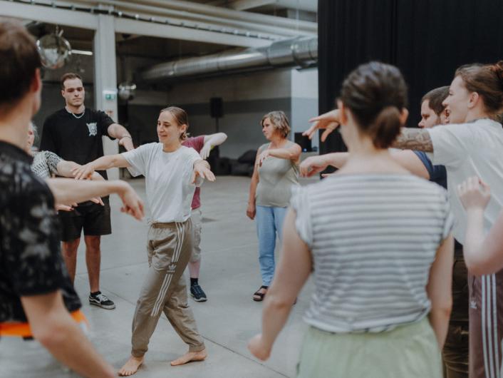 Eine Gruppe von Menschen steht im Kreis und macht bei einem Bewegungs- oder Tanzworkshop in einem großen, offenen Raum mit. Die Stimmung ist freundlich und ungezwungen.
