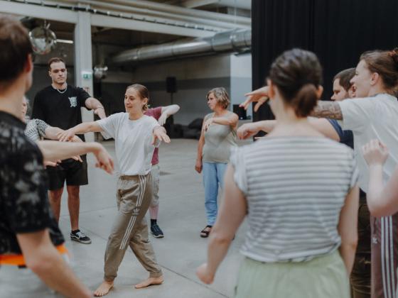 A group of people is standing in a circle, taking part in a movement or dance workshop in a large, open room. The atmosphere is friendly and informal.