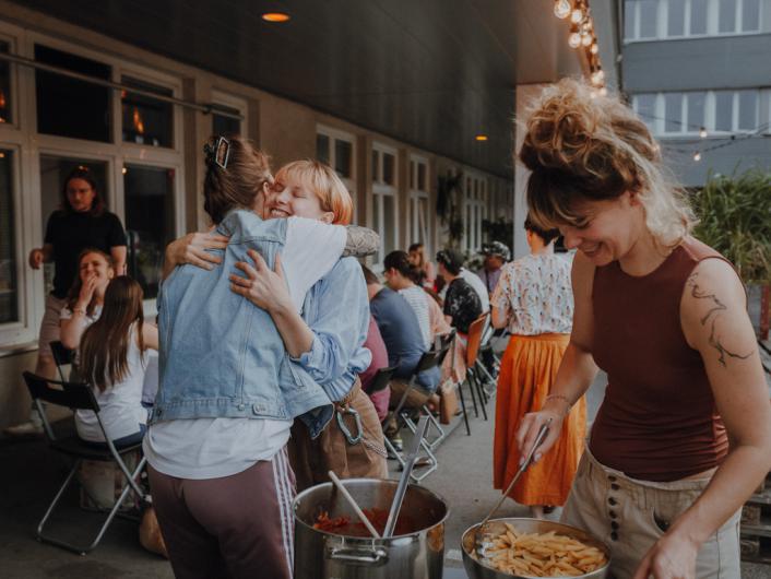 Menschen sitzen draußen auf einer überdachten Terrasse und essen gemeinsam. Im Vordergrund umarmen sich zwei Personen herzlich, während eine andere Person mit einem Lächeln Essen aus einem großen Topf serviert.