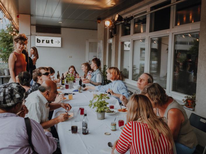 Eine lange Tafel auf einer Terrasse, an der eine vielfältige Gruppe von Menschen zusammen isst, spricht und das Beisammensein genießt. An der Wand hängt ein Schild mit der Aufschrift „brut“.