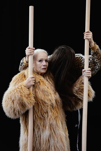 Zwei Frauen, eine mit langen weißen Haaren und einem beigen Pelzmantel, die andere mit braunen Haaren und einer Jacke mit Leopardenmuster, stehen dicht beieinander und halten Holzstangen vor einem schwarzen Hintergrund.