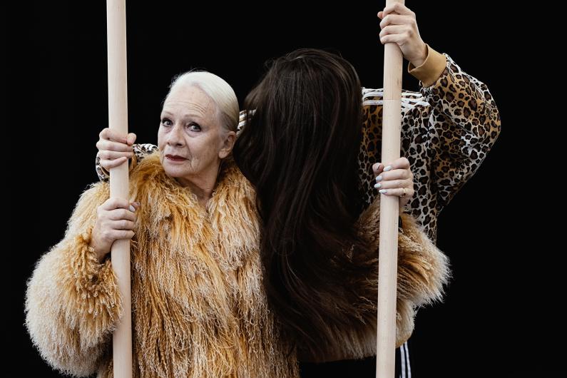 Zwei Frauen, eine mit langen weißen Haaren und einem beigen Pelzmantel, die andere mit braunen Haaren und einer Jacke mit Leopardenmuster, stehen dicht beieinander und halten Holzstangen vor einem schwarzen Hintergrund.