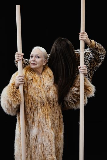 Zwei Frauen, eine mit langen weißen Haaren und einem beigen Pelzmantel, die andere mit braunen Haaren und einer Jacke mit Leopardenmuster, stehen dicht beieinander und halten Holzstangen vor einem schwarzen Hintergrund.