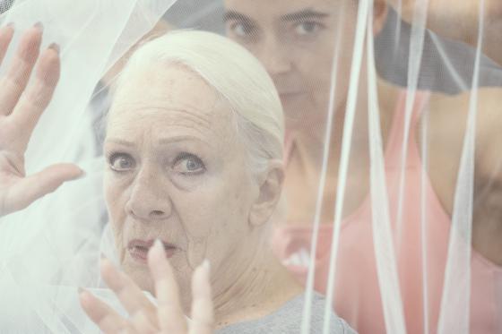 Eine Nahaufnahme einer älteren Frau mit weißem Haar und ernstem Ausdruck, teilweise durch weißen Tüll verschleiert. Hinter ihr blickt eine jüngere Person in einem rosa Oberteil, deren Gesicht teilweise durch den Stoff sichtbar ist.