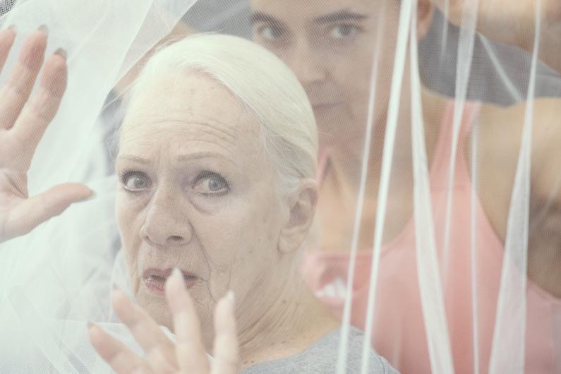 Eine Nahaufnahme einer älteren Frau mit weißem Haar und ernstem Ausdruck, teilweise durch weißen Tüll verschleiert. Hinter ihr blickt eine jüngere Person in einem rosa Oberteil, deren Gesicht teilweise durch den Stoff sichtbar ist.